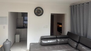 Uma sala de estar moderna com um sofá de couro, um relógio na parede e uma cozinha visível através de uma porta aberta.