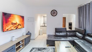 Uma sala de estar moderna com sofá de couro, televisão de tela plana, mesa de centro de madeira e cozinha ao fundo.