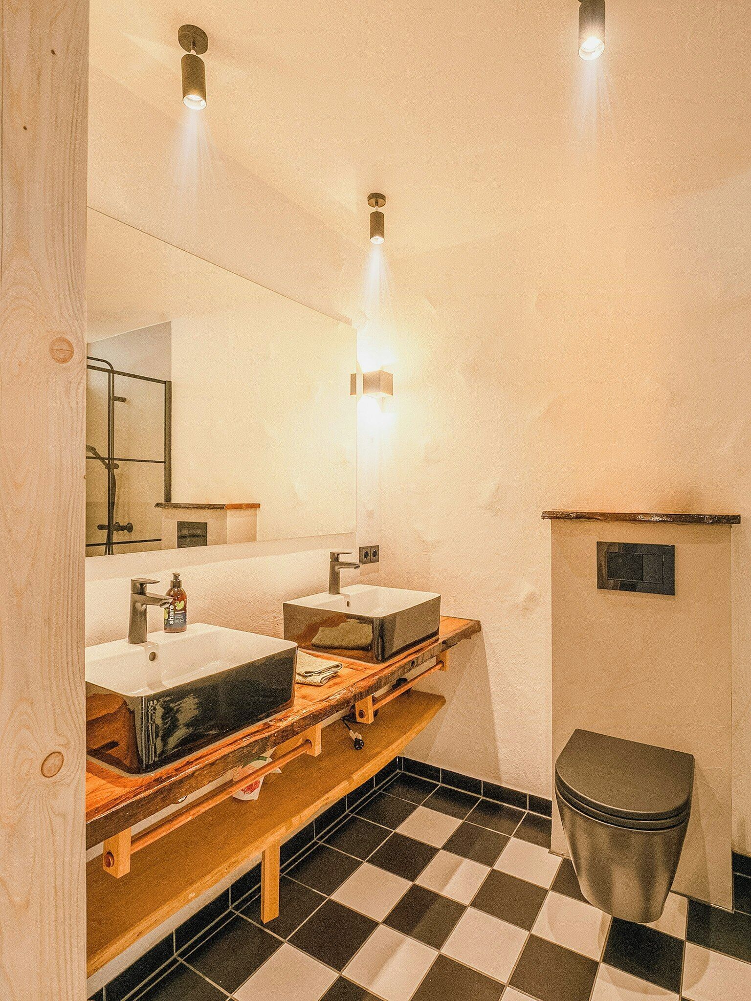 Une salle de bain avec deux lavabos, un miroir, une étagère en bois, une poubelle et un carrelage à motifs carrés.