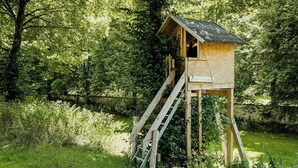 A wooden treehouse with a ladder, surrounded by dense greenery and a small stream. - Experience original nature in the steward's house (Kall)