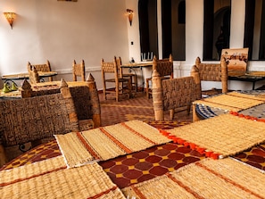 A room with woven furniture and rugs, a dining area with tables and chairs, and a view of the outside through windows. - Riad Bleu Afriqua (Ait Sedrate Jbel El Soufla)