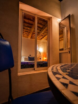 A room with a bed, a window with a view of a room with a lamp, and a table with a patterned tablecloth. - Riad Bleu Afriqua (Ait Sedrate Jbel El Soufla)