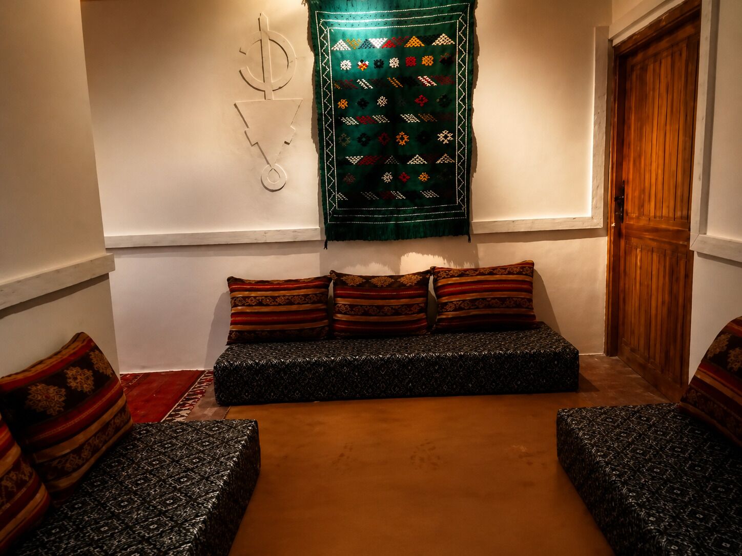 A cozy room with a sofa set, patterned cushions, a decorative wall hanging, and a wooden door.