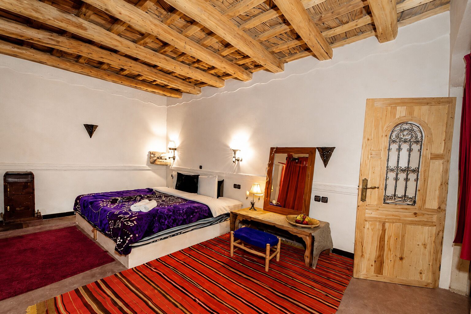 A room with a bed, a desk, a chair, a wooden door, and a red carpet.