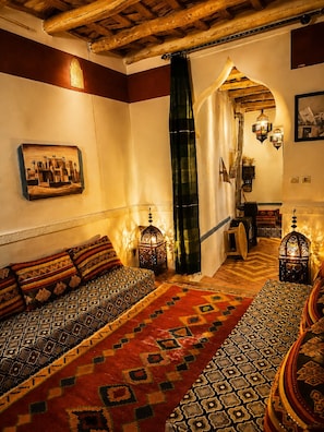 A traditional Moroccan interior with a wooden ceiling, a patterned rug, a sofa with cushions, and wall-mounted lanterns.