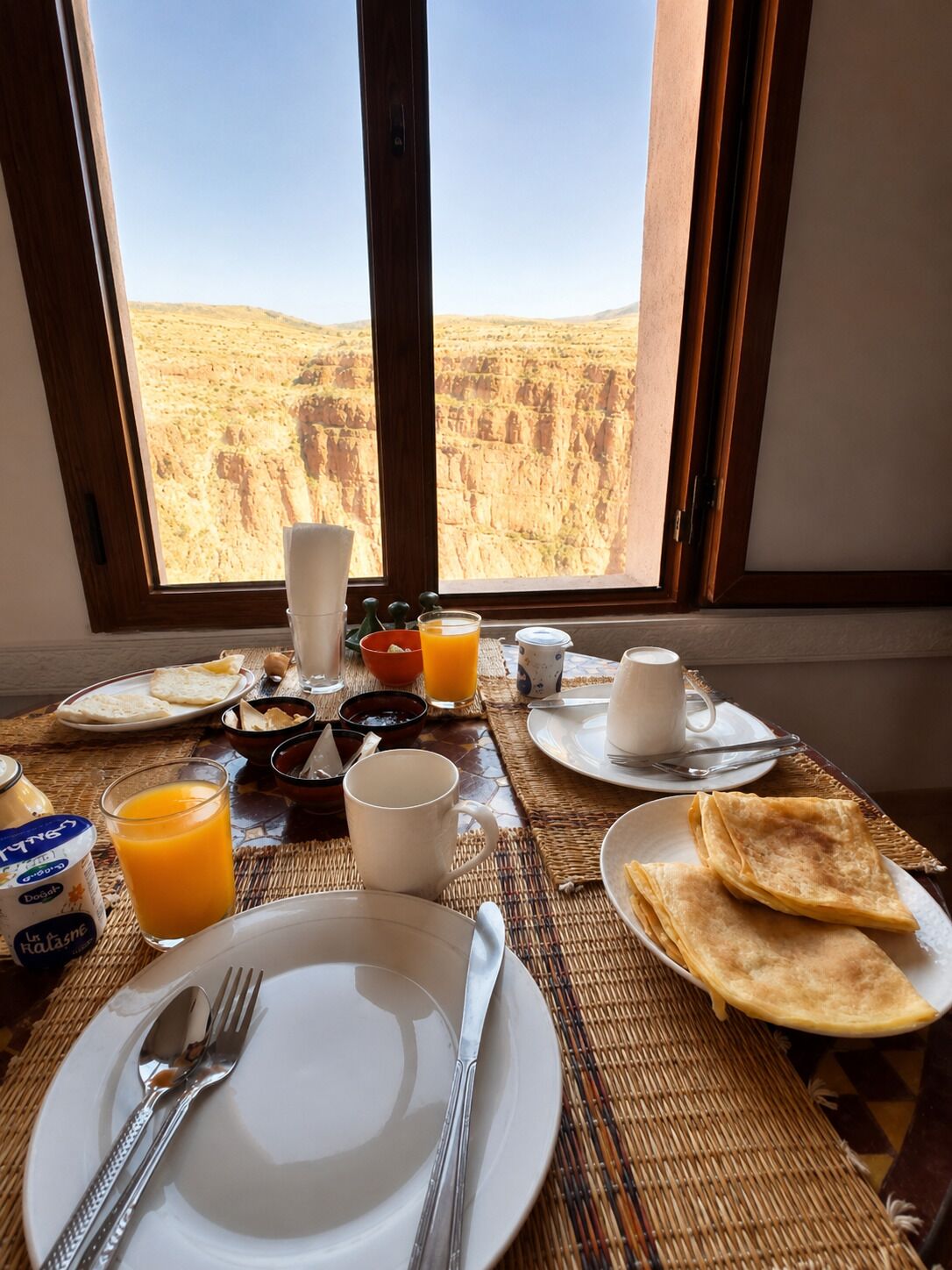 A breakfast table with plates, cutlery, and a glass of orange juice, set against a view of a desert landscape.