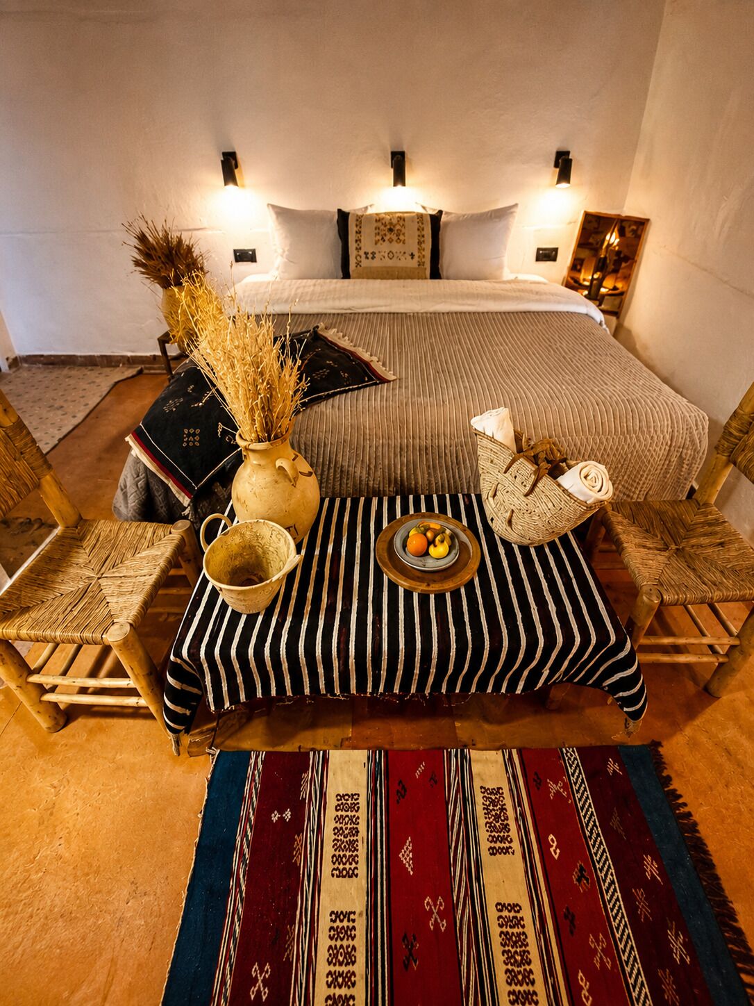 A room with a bed, two chairs, a table with a bowl of fruit, and decorative pillows.