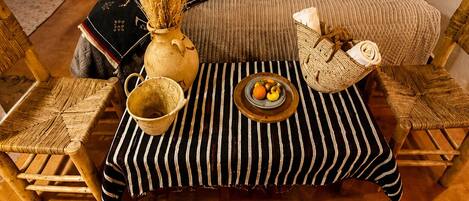 A room with a bed, two chairs, a table with a bowl of fruit, and decorative pillows.