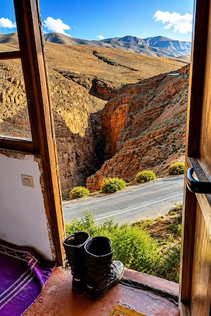 A window view of a mountainous landscape with a road, a purple cloth, and black boots on a wooden ledge. - Riad Bleu Afriqua (Ait Sedrate Jbel El Soufla)