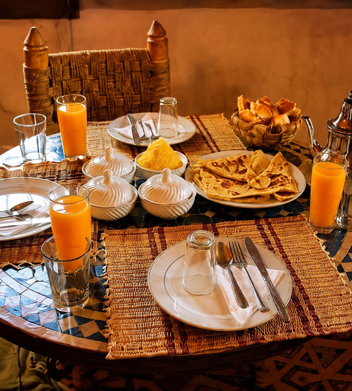 A breakfast setting with a table set for two, including plates, glasses, and a basket of bread.