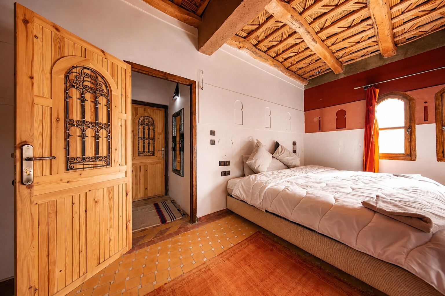 A bedroom with a wooden door, a bed with white bedding, a window with curtains, and a tiled floor.