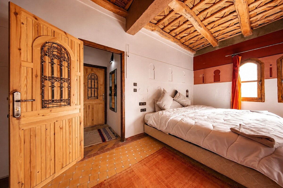 A bedroom with a wooden door, a bed with white bedding, a window with curtains, and a tiled floor.