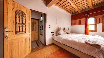 A bedroom with a wooden door, a bed with white bedding, a window with curtains, and a tiled floor.