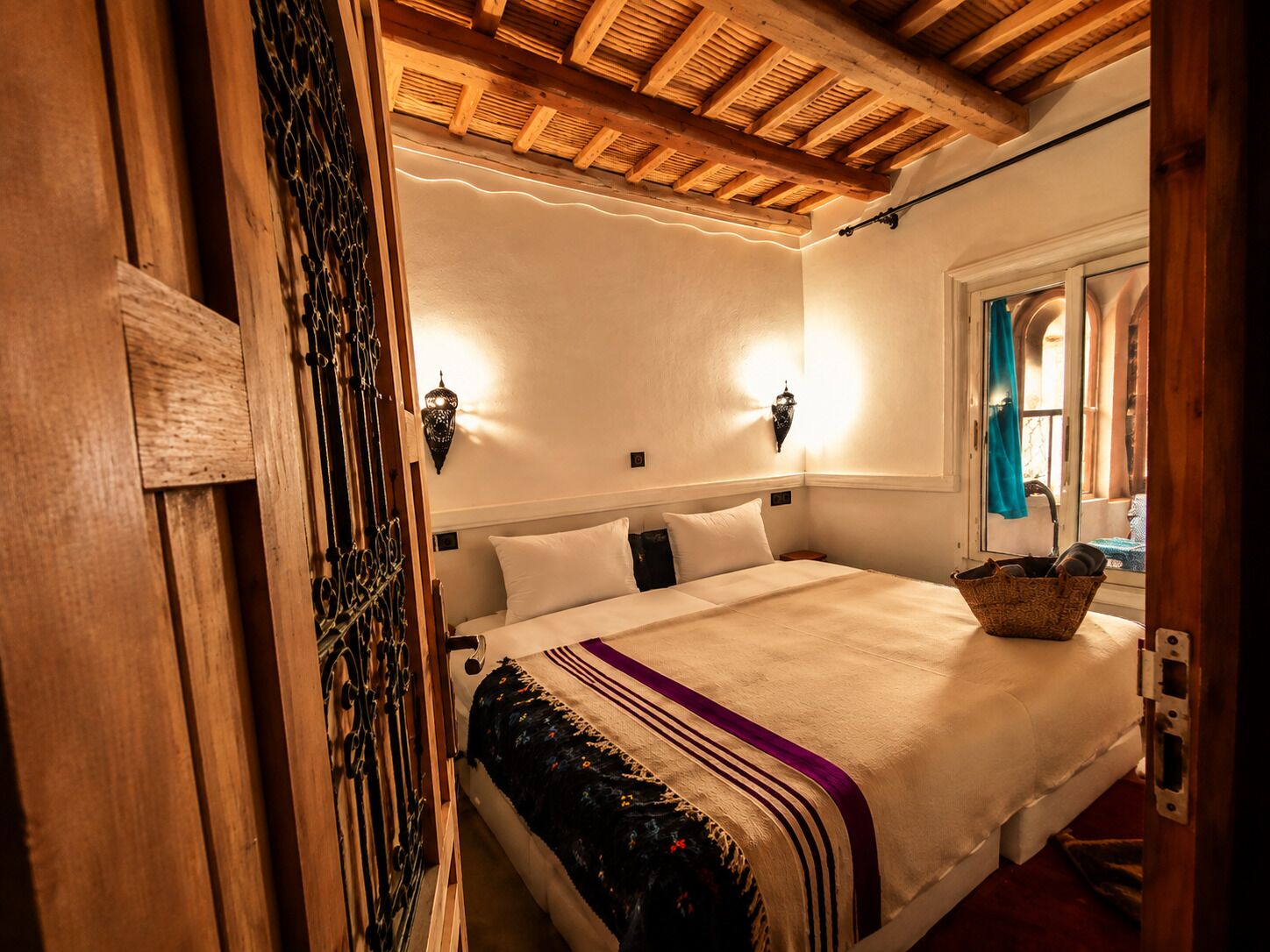 Una camera da letto con soffitto in legno, un letto con copripiumino bianco e viola, due lampade a parete e una finestra con vista esterna.