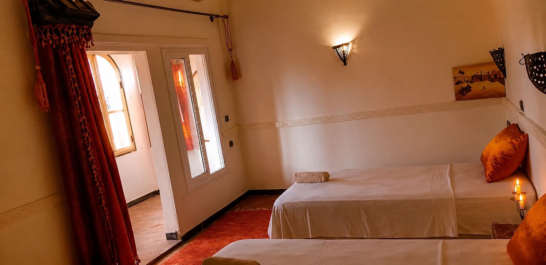 Quarto de hotel com duas camas, um tapete vermelho, luminária na parede e um quadro.