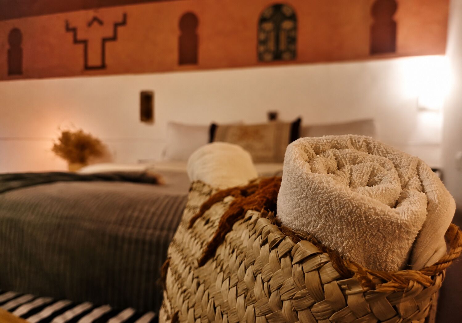A bed with a woven basket containing towels, a wall with geometric patterns, and a bedside table with a vase.