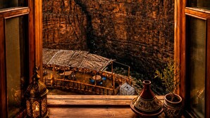 A view through a window of a mountainous landscape at sunset, with a small outdoor seating area visible below. - Riad Bleu Afriqua (Ait Sedrate Jbel El Soufla)