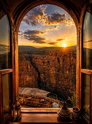 A view through a window of a mountainous landscape at sunset, with a small outdoor seating area visible below.