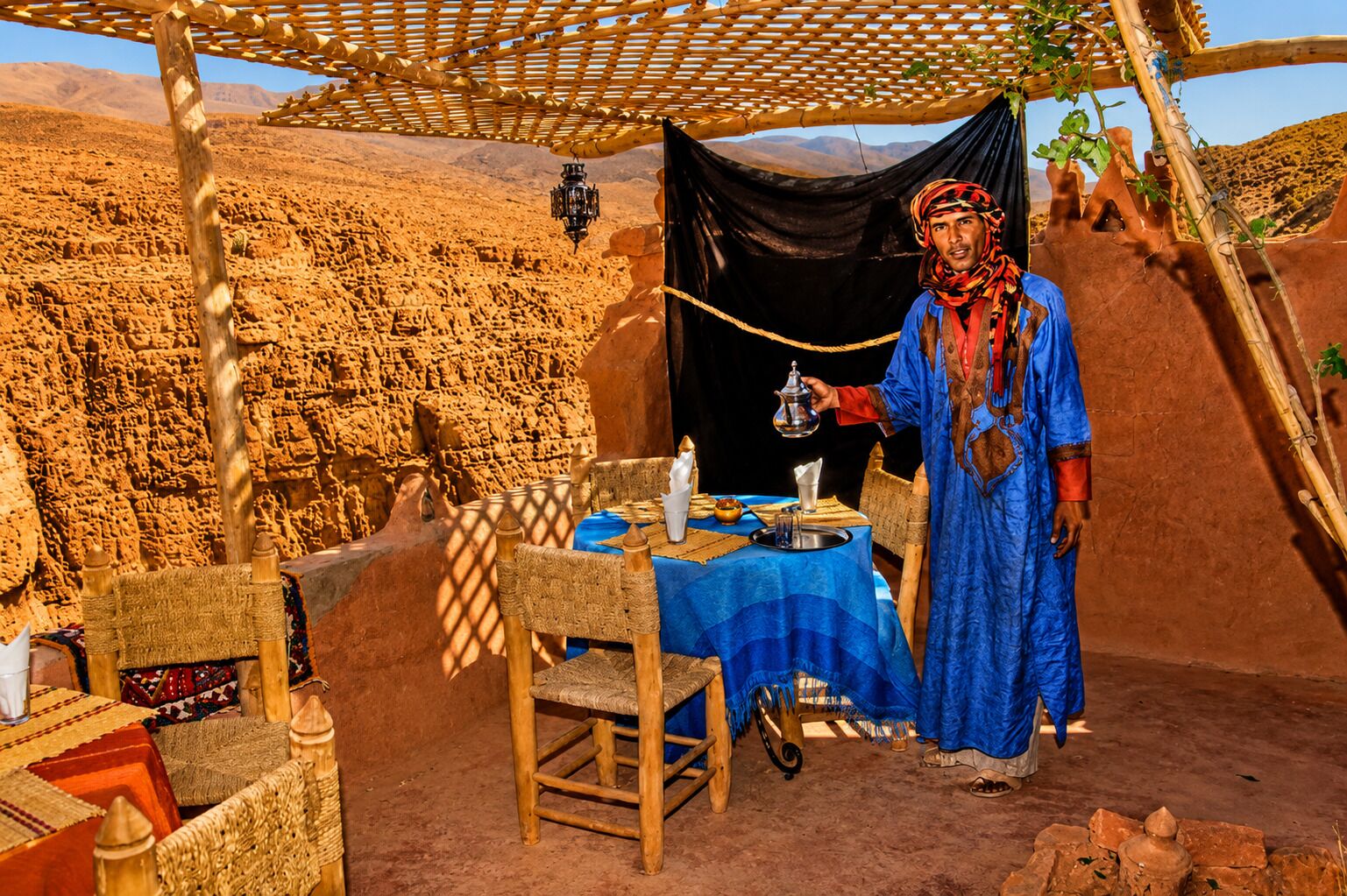 A desert tent with a table set for a meal, a person serving, and a mountainous backdrop.