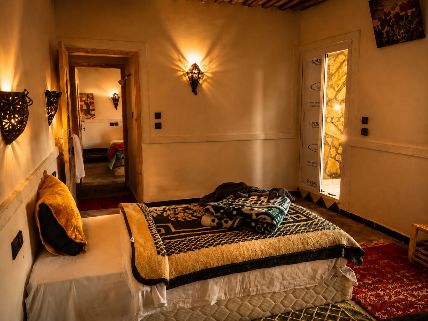 Habitación de hotel con cama, alfombra roja, puerta con diseño de piedra y luces fijadas a la pared.