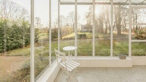 Ein Wintergarten mit weißem Stuhl und Tisch, großen Fenstern und Blick auf einen Garten.