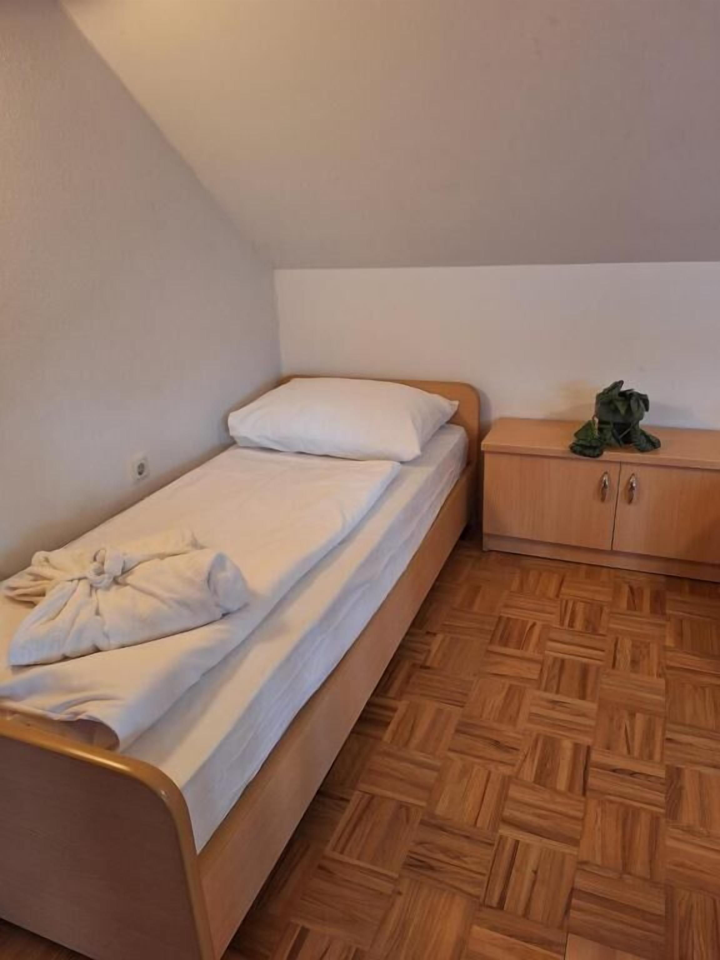 A single bed with a wooden headboard, a small wooden nightstand with a green object on it, and a herringbone-patterned wooden floor.