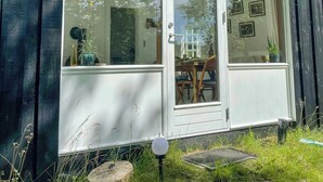 Un edificio de madera con un gran escaparate y ventanas, un pequeño espacio al aire libre con mesas y sillas, y un timbre visible.
