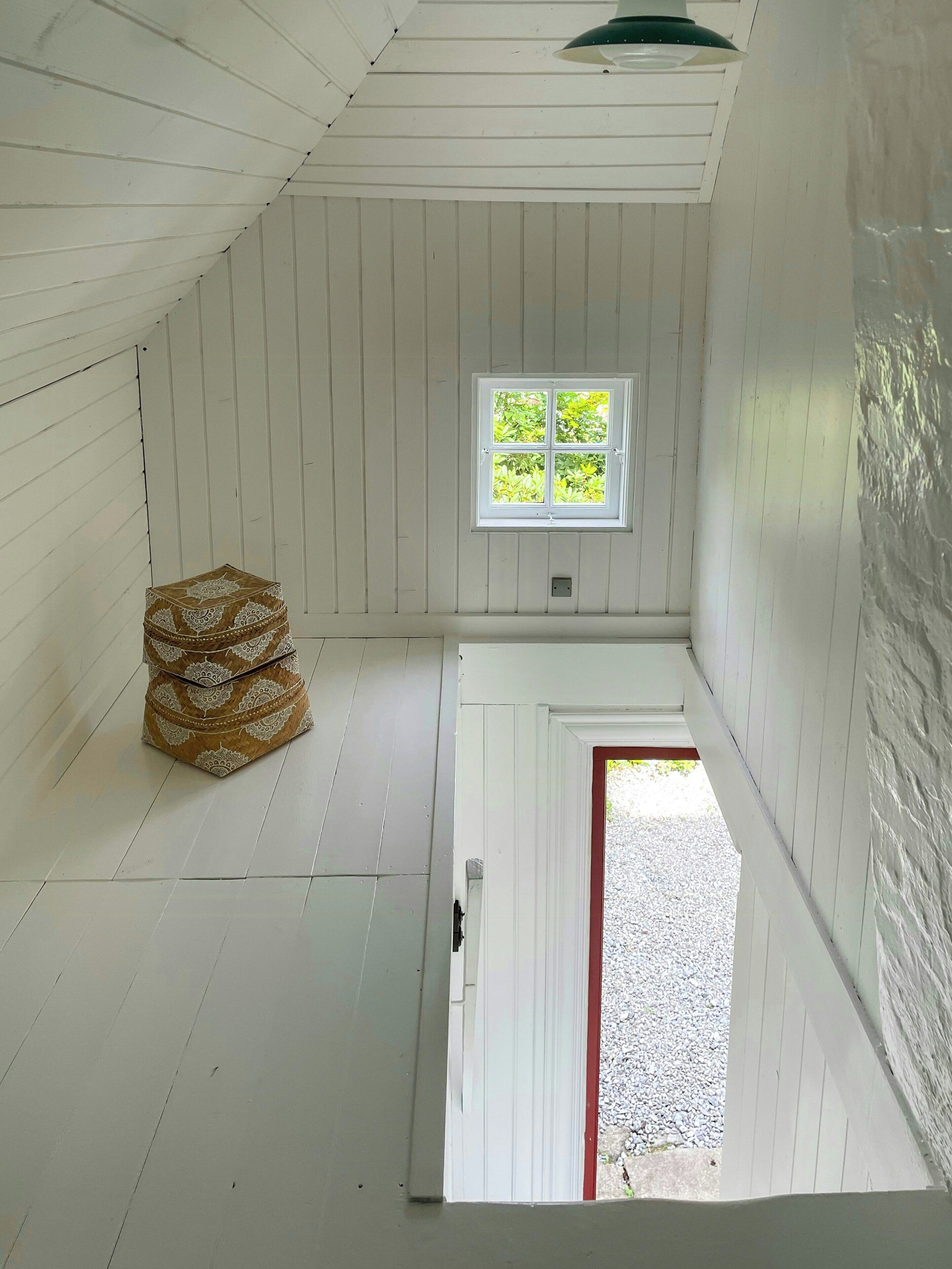 A room with white wooden walls, a window with a view of greenery, a patterned ottoman, and a door leading outside.