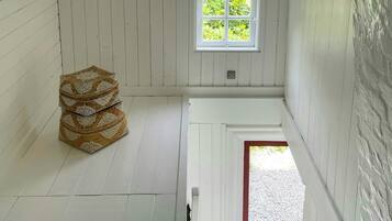 A room with white wooden walls, a window with a view of greenery, a patterned ottoman, and a door leading outside.