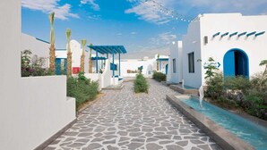 A pathway leading to a pool area with white buildings and blue accents, surrounded by greenery.