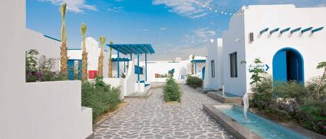 A pathway leading to a pool area with white buildings and blue accents, surrounded by greenery.