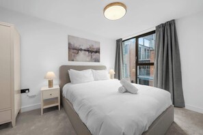 Un dormitorio con cama, mesitas de noche, un cuadro y una ventana con cortinas.