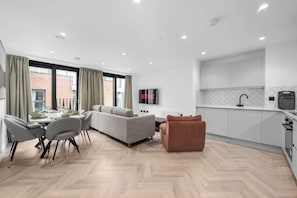 Un salon moderne comprenant un coin repas, un canapé, un pouf marron, une cuisine avec des meubles blancs et un téléviseur fixé au mur.