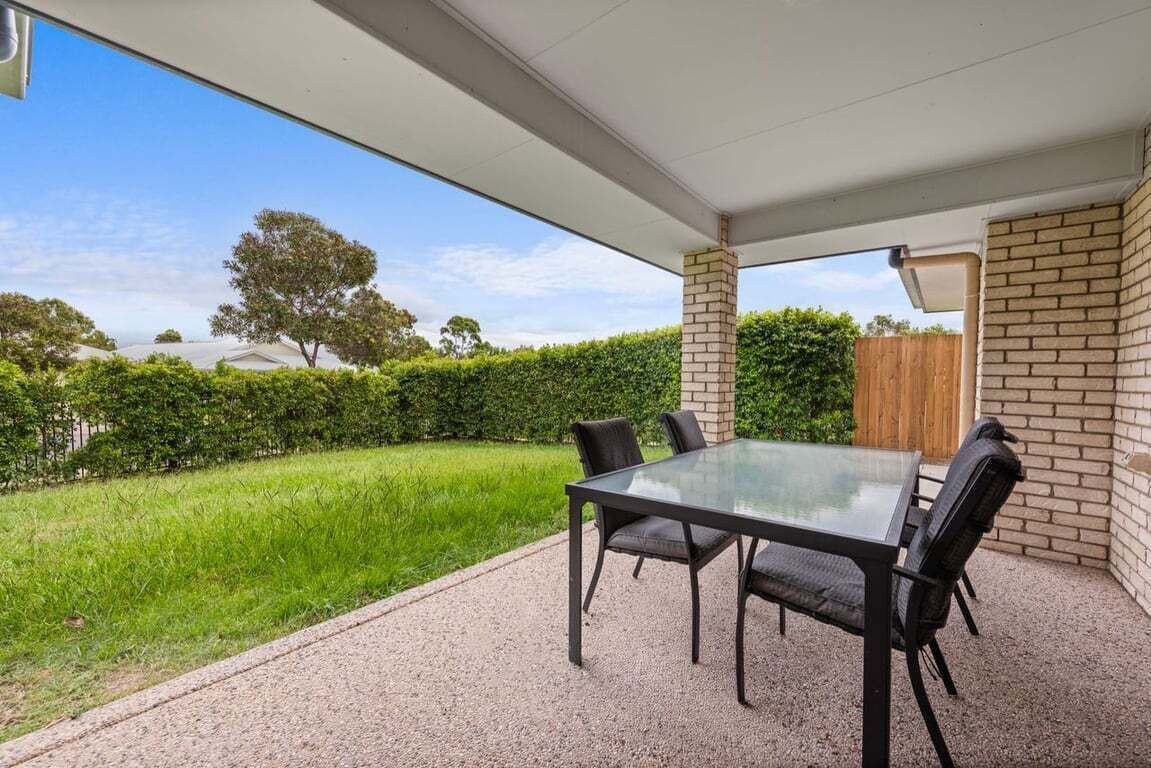 A covered patio with a glass table and chairs, a brick wall, and a view of a green lawn and trees.