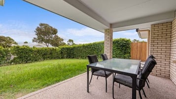 A covered patio with a glass table and chairs, a brick wall, and a view of a green lawn and trees.