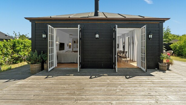 Un edificio de madera con fachada oscura, puertas abiertas que dan a un interior con cama, mesa y sillas.