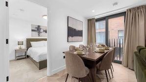Et hotelværelse med spiseplads, seng, sengebord, lampe og et vindue med gardiner.