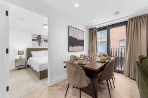 Habitación de hotel con comedor, cama, mesita de noche, lámpara y ventana con cortinas.