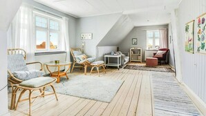 Een gezellige kamer onder de dakspanten, met houten vloeren en diverse zitmogelijkheden, waaronder rieten stoelen en een rode fauteuil.