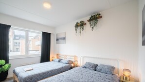 Quarto de hotel com duas camas, uma janela, um radiador, uma planta suspensa na parede e um abajur.