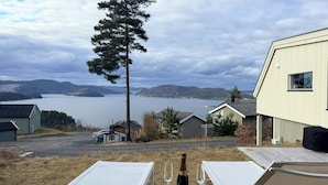 Blick auf einen See mit einem Berg im Hintergrund, einen hohen Baum und ein Gebäude mit weißer Fassade.