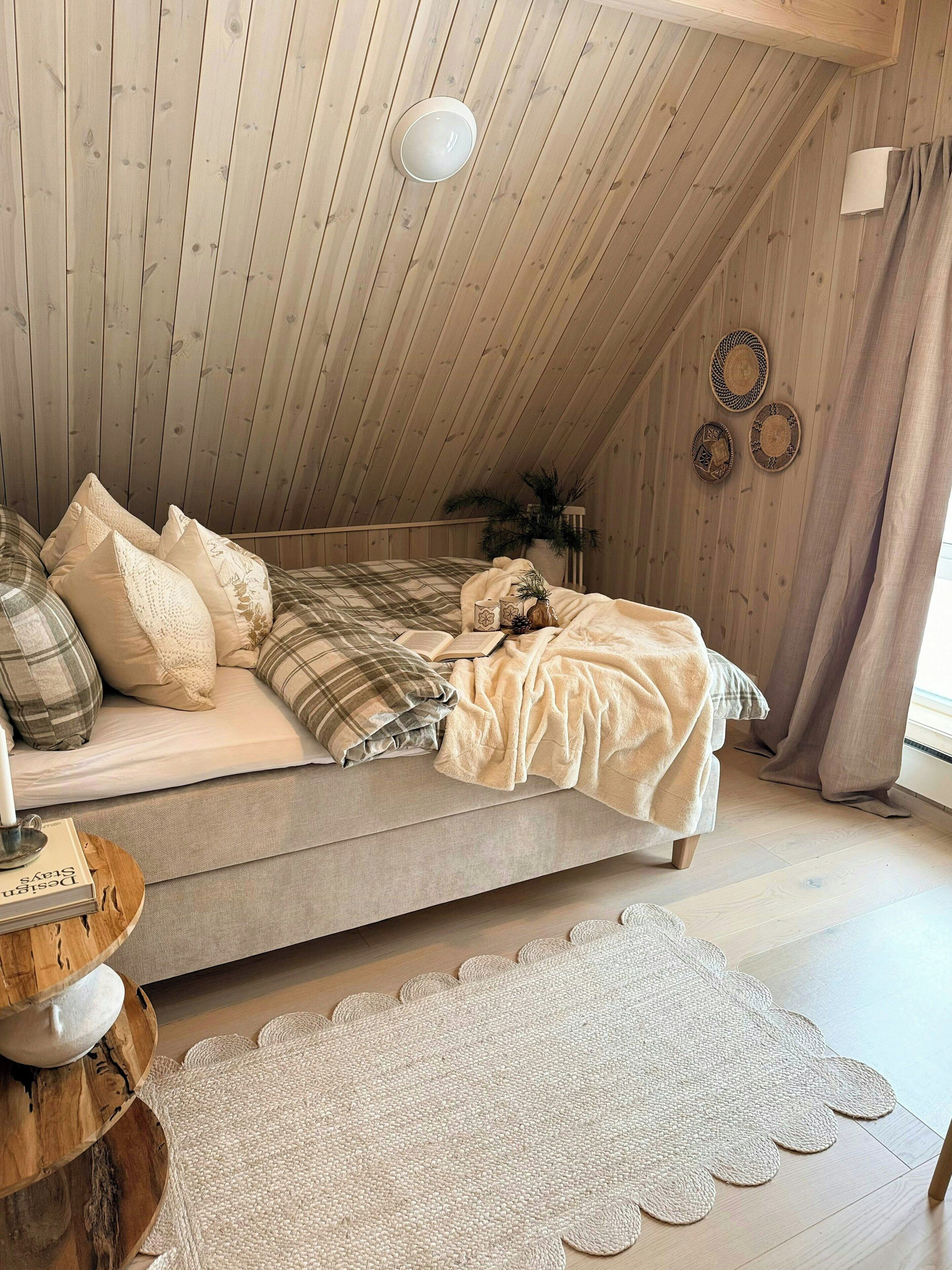 Ein gemütliches Schlafzimmer mit schrägem Dach, einem Bett mit Karo-Bettwäsche, einem Nachttisch mit Buch und einer Wanddekoration.