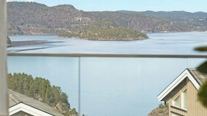 Ein See mit bewaldeten Hügeln im Hintergrund, ein Haus mit Balkon und einem klaren Himmel.