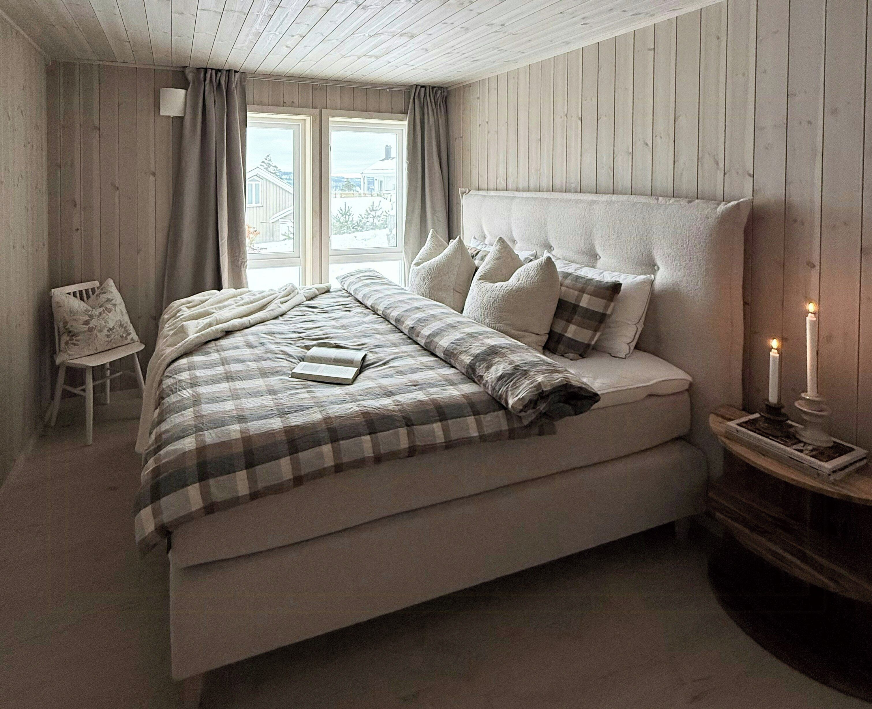 A bedroom with a bed, a chair, a bedside table with candles, and a window with curtains.