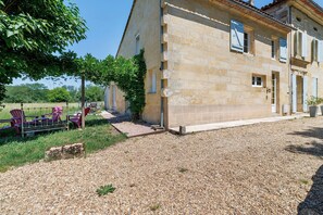 Un bâtiment en pierre avec une allée gravillonnée, des sièges à l’extérieur et un arbre qui offre de l’ombre.