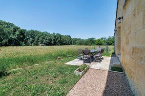 Eine Terrasse im Freien mit Tisch und Stühlen, ein Kiesweg und ein Grasfeld mit Bäumen im Hintergrund.