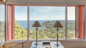 A room with a large window offering a scenic view of a coastal landscape, a table with two lamps, and a clock on the wall. - Villa with Pool and Spectacular Sea Views (Begur)