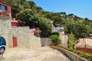 Edifici amb parets de pedra, una porta vermella i un cotxe blau a la façana, amb vistes a un turó ple d'arbres i un cel serè.