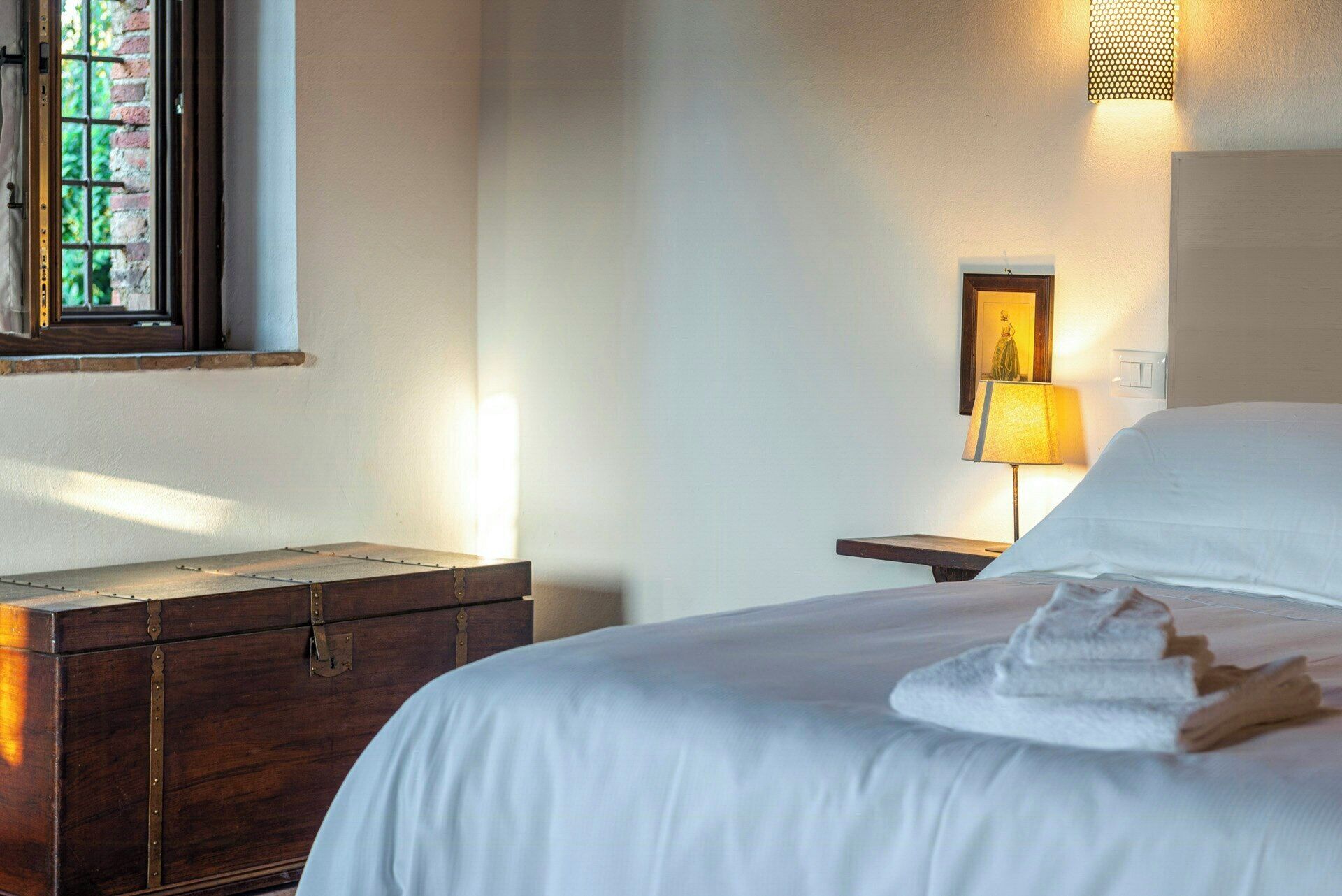 Une chambre d’hôtel avec un lit, une commode en bois, une lampe et une fenêtre donnant sur un paysage verdoyant.
