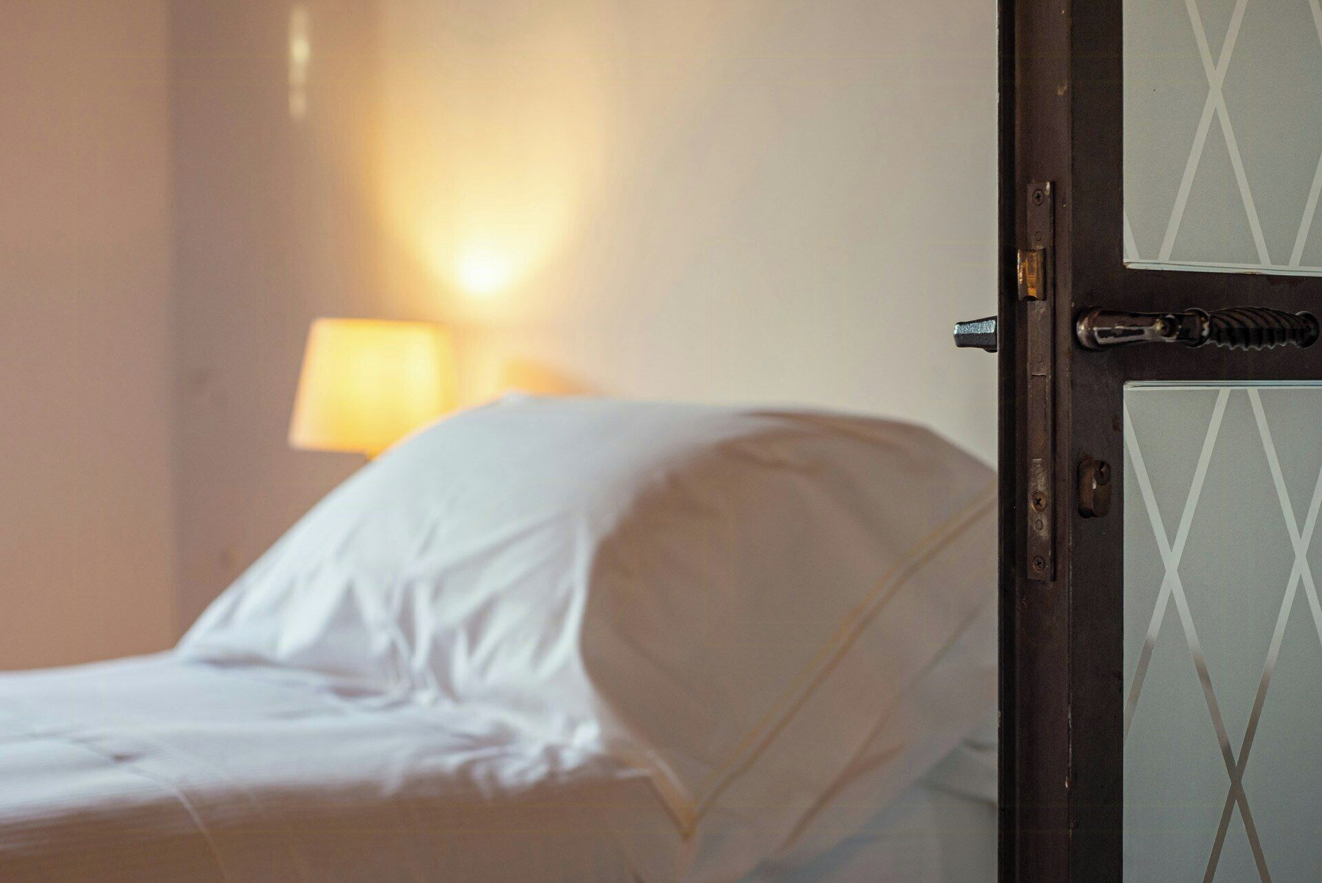 A bed with white bedding, a door with a metal handle, and a wall-mounted lamp.